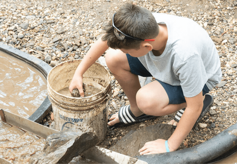 A boy dips a muddy rock into a bucket of water at Big Thunder Gold Mine while gold panning