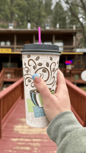 A close-up of a person holding a large hot coffee cup with a coffee bean pattern while standing on a wooden bridge.