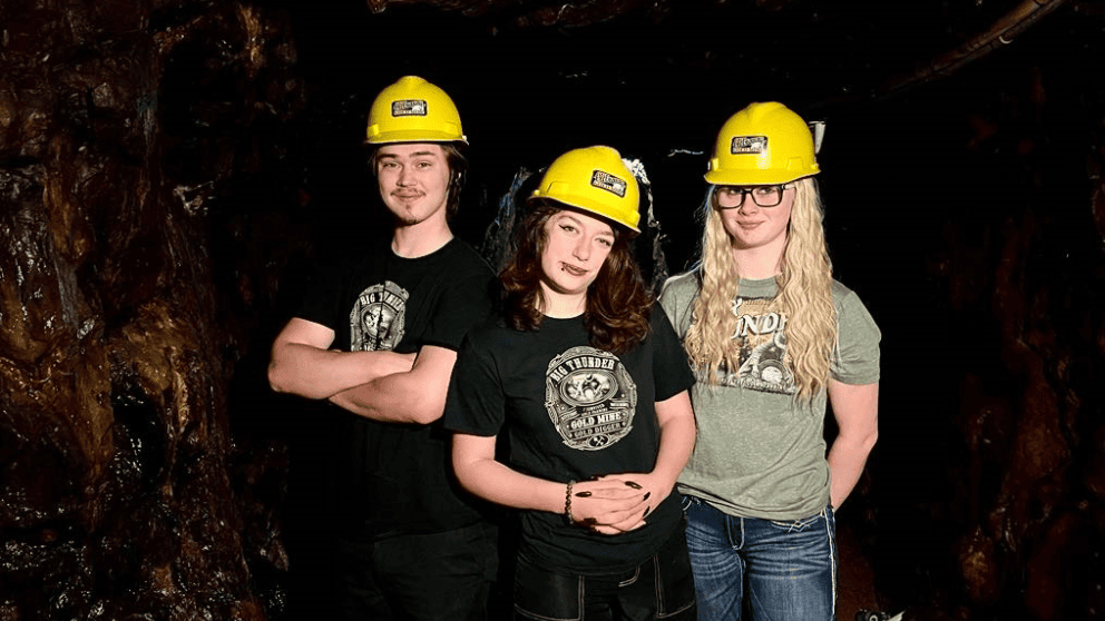 Three young adults wearing Big Thunder Gold Mine yellow hard hats and graphic t-shirts in a mine shaft
