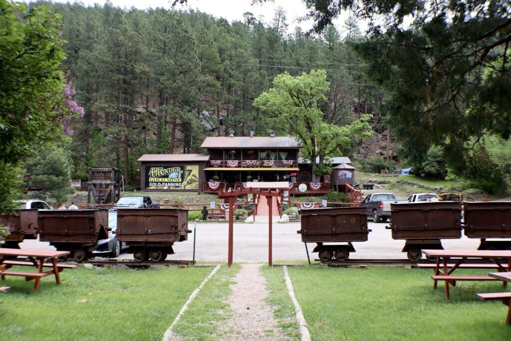 Exterior view of the Big Thunder Gold Mine in Keystone, South Dakota, showing historic mining carts, an outdoor picnic area, and the main log cabin visitor center against a forested hill.